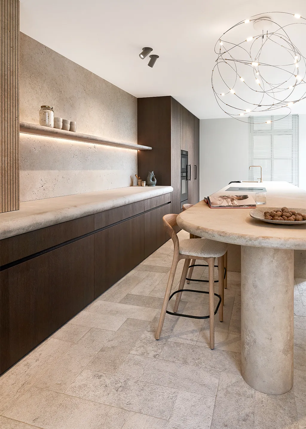 Luxe Japandi keuken van Eggersmann met greploze kasten in warm bruin hout, organisch afgerond kookeiland met travertin natuursteen werkblad en minimalistische houten wandplank